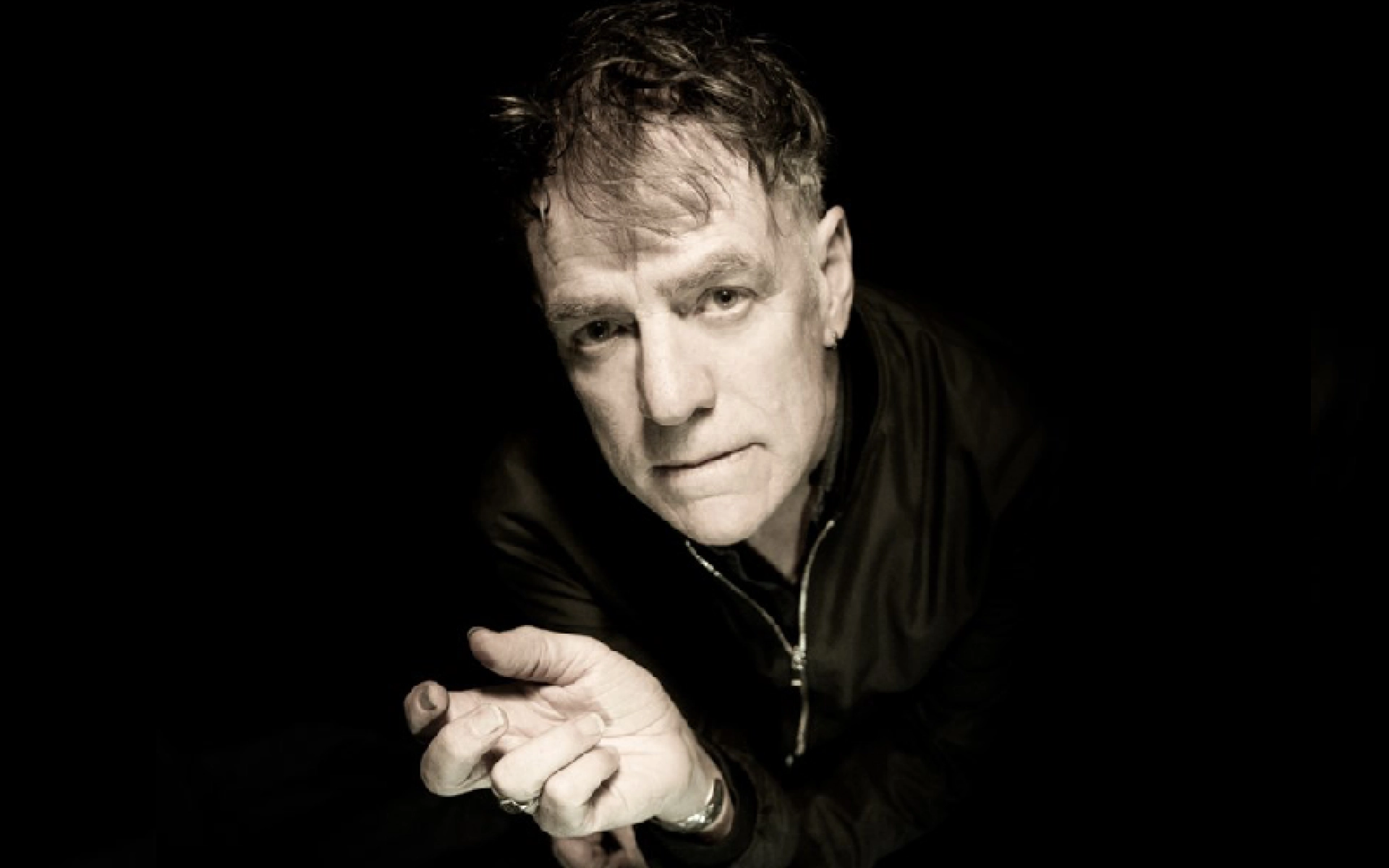 Portrait of a person with short hair and stubble, leaning toward the camera, hands clasped, wearing a dark jacket, against a black background.