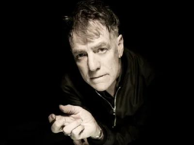 Portrait of a person with short hair and stubble, leaning toward the camera, hands clasped, wearing a dark jacket, against a black background.