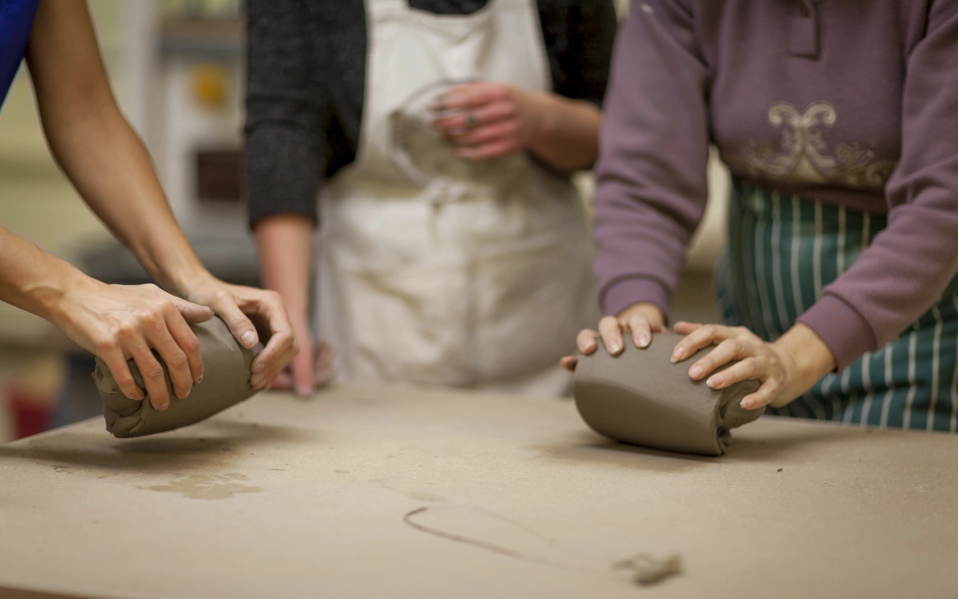 An image of three people in a pottery studio, their faces out of view with a focus on two peoples hands kneading clay at a table.