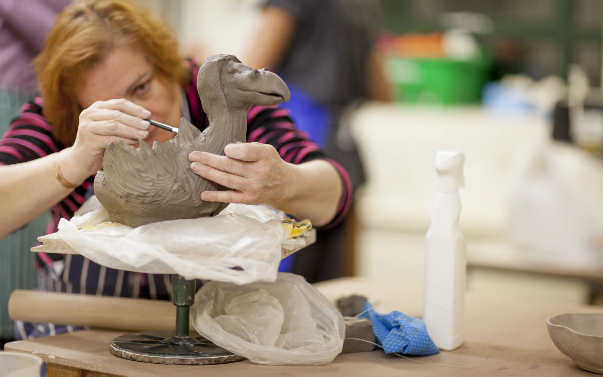 Set in a pottery studio, a woman delicately carves details onto a large clay bird sculpture with it in the foreground.