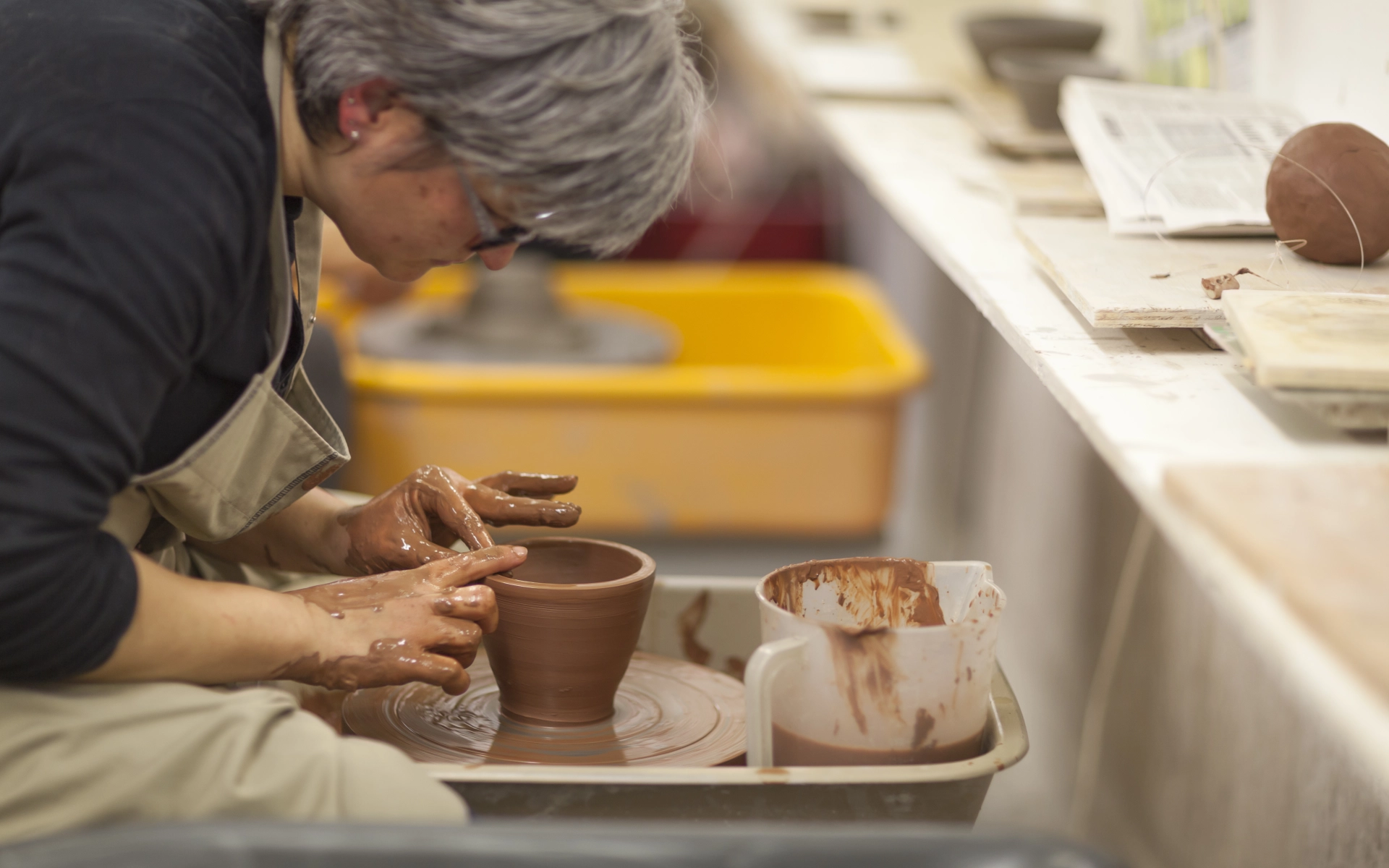 A person is sat at a pottery throwing wheel in a studio. They have their hands on the clay, sculpting it into a bowl-like shape.