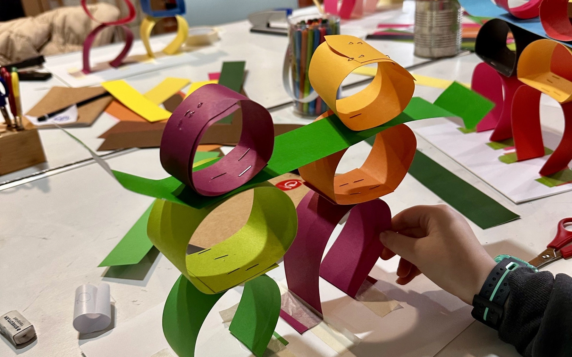 Two small figures stand on an arts and crafts table, the figures are made from circles of colourful paper and held together with staples. A child's hand holds them upright. In the background more of these figures can be seen on the table.