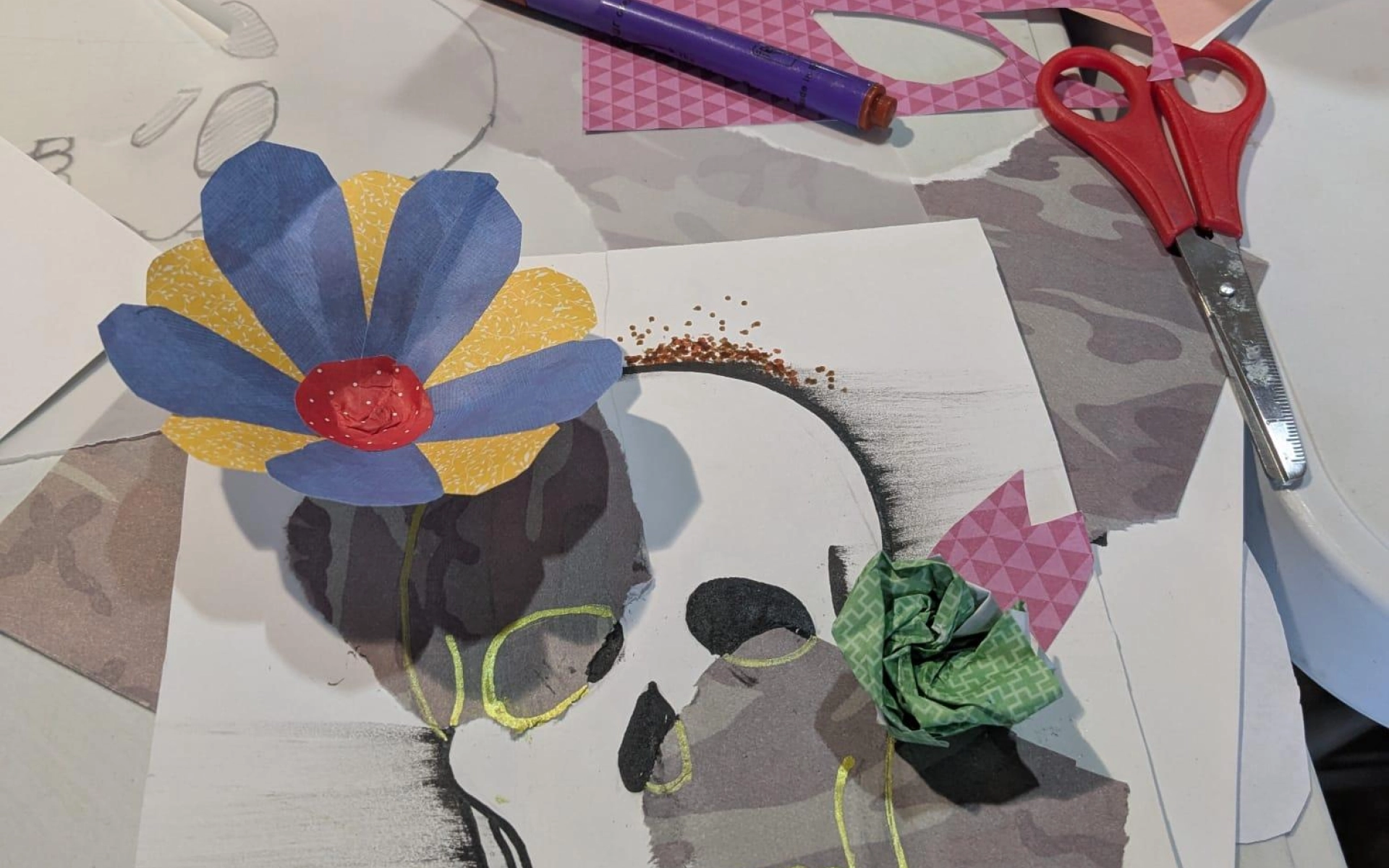 An image of a skull made up of drawings and paper collage. It has a blue and yellow paper flower on top. The drawing is on a table of craft pieces, pens and scissors.