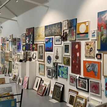 A bright art gallery with white walls and ceiling beams, densely packed framed paintings of landscapes, abstracts and portraits.
