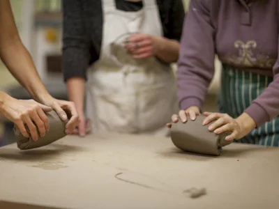 An image of three people in a pottery studio, their faces out of view with a focus on two peoples hands kneading clay at a table.