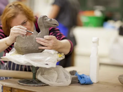 Set in a pottery studio, a woman delicately carves details onto a large clay bird sculpture with it in the foreground.