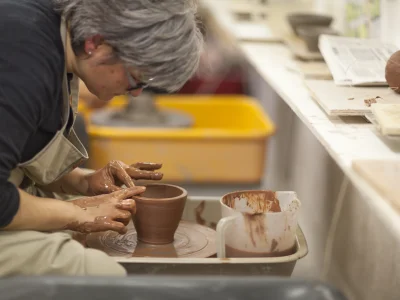 A person is sat at a pottery throwing wheel in a studio. They have their hands on the clay, sculpting it into a bowl-like shape.