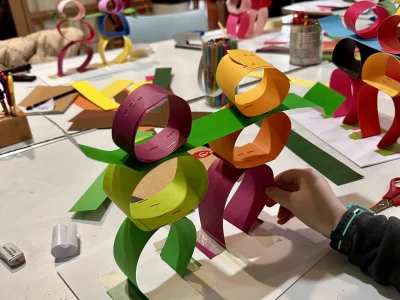 Two small figures stand on an arts and crafts table, the figures are made from circles of colourful paper and held together with staples. A child's hand holds them upright. In the background more of these figures can be seen on the table.