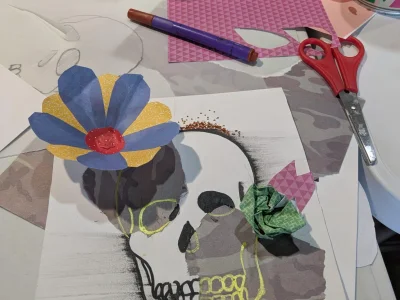 An image of a skull made up of drawings and paper collage. It has a blue and yellow paper flower on top. The drawing is on a table of craft pieces, pens and scissors.