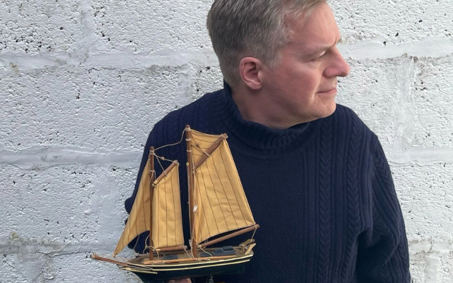 Person in a dark blue knitted jumper holds a small wooden sailing ship model with tan sails, against a pale brick wall.