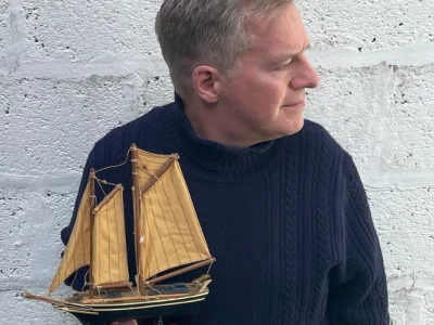 Person in a dark blue knitted jumper holds a small wooden sailing ship model with tan sails, against a pale brick wall.