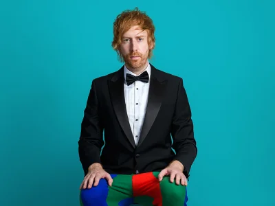 Person with ginger hair and beard in a black tuxedo and bow tie sits against a teal background, hands on bright patchwork trousers in blue, green and red.