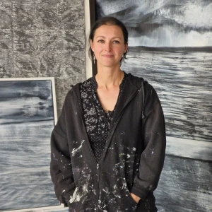 Portrait of a person with dark hair in a paint-splattered black hoodie, smiling, standing in an art studio with large monochrome ocean paintings.