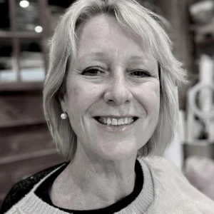 Smiling person with short blonde hair and a pearl earring, wearing a light knit sweater; softly blurred indoor background.