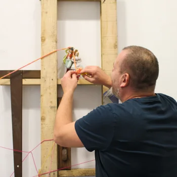An adult in a navy T‑shirt adjusts a small figurine mounted on a wooden frame, with pink strings stretched across a white wall backdrop.