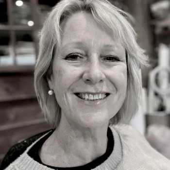 Smiling person with short blonde hair and a pearl earring, wearing a light knit sweater; softly blurred indoor background.