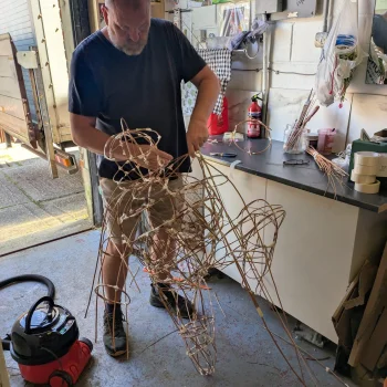 A person in a workshop crafts a large, tangled wire sculpture near a workbench with tools, shelves and a red fire extinguisher.