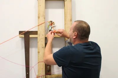 An adult in a navy T‑shirt adjusts a small figurine mounted on a wooden frame, with pink strings stretched across a white wall backdrop.