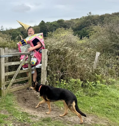 Person wearing a pink cardboard costume with a yellow cone hat stands at a wooden gate, while a German Shepherd walks along a dirt path beside tall hedges.