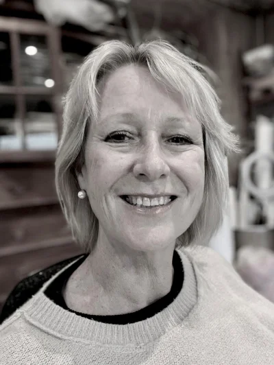 Smiling person with short blonde hair and a pearl earring, wearing a light knit sweater; softly blurred indoor background.