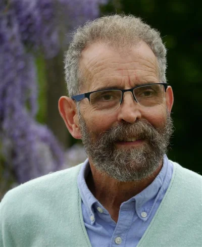 A person with a grey beard and glasses, smiling slightly, wearing a light blue shirt and mint cardigan, outdoors with purple flowers in the background.