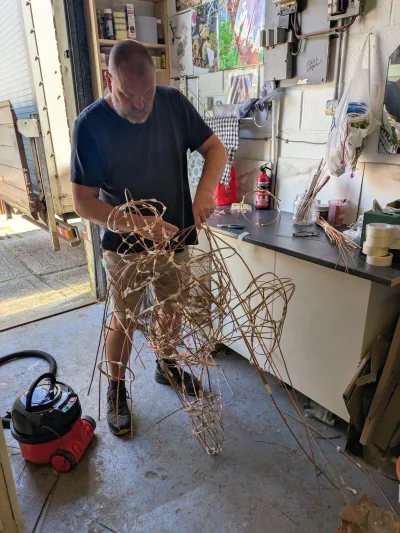 A person in a workshop crafts a large, tangled wire sculpture near a workbench with tools, shelves and a red fire extinguisher.