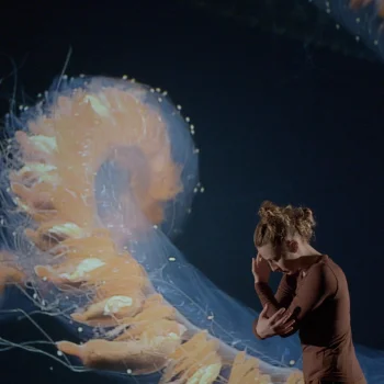 A performer in brown clothing stands in a dark setting beside a large translucent orange jellyfish-like sculpture with dangling tendrils.