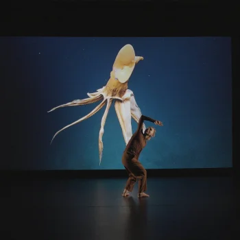 A dancer in brown clothes performs on a dark stage before a large screen showing a beige squid with long tentacles against a blue backdrop.