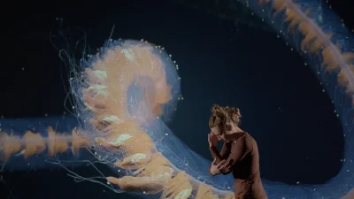 A performer in brown clothing stands in a dark setting beside a large translucent orange jellyfish-like sculpture with dangling tendrils.