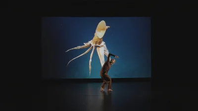 A dancer in brown clothes performs on a dark stage before a large screen showing a beige squid with long tentacles against a blue backdrop.