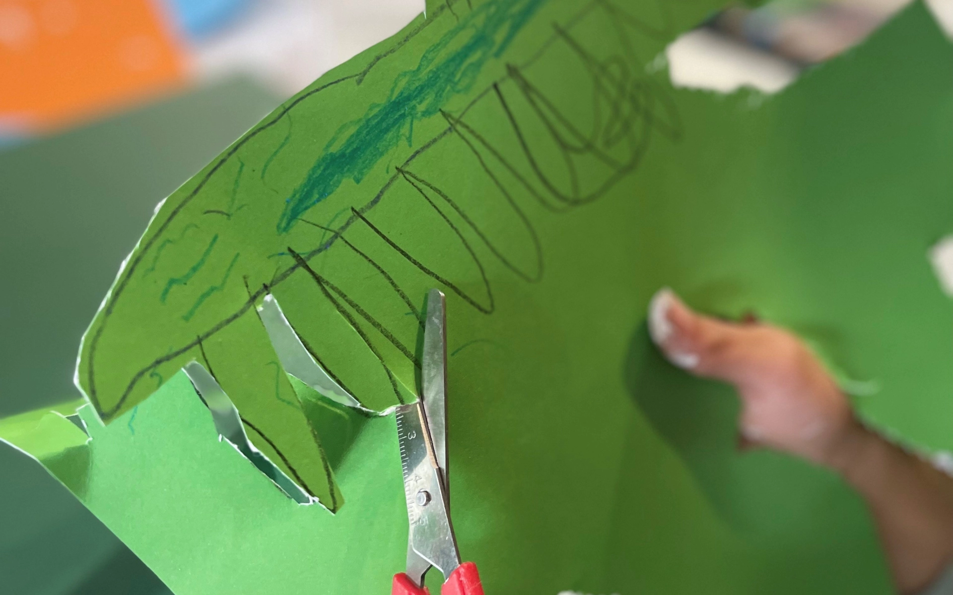 An image featuring a child's hands, they are cutting a green piece of card with scissors in their left hand. The card has a sea creature drawn on.