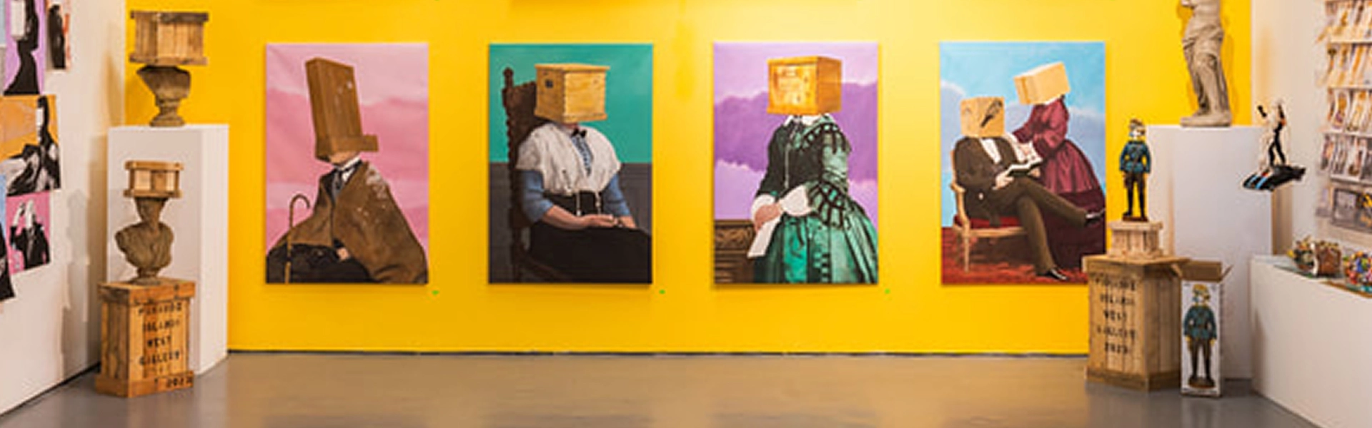 Art gallery with a yellow wall and five portraits of people with boxes on their heads; pedestals hold sculptures and magazines line the side.