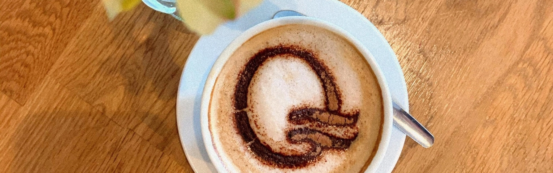 Top-down view of a white coffee cup on a light blue saucer, on a wooden table; cocoa powder forms a G-shaped design on the frothy surface with a spoon nearby.