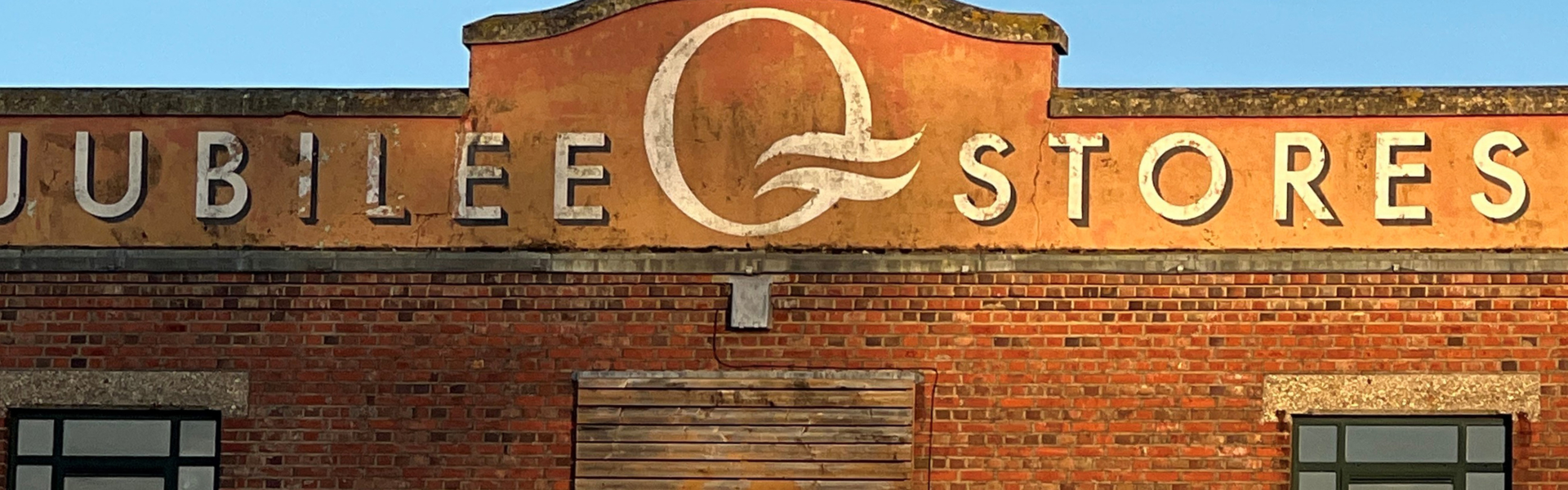 Brick storefront with an orange, weathered sign reading JUBILEE STORES in white letters and a central circular motif, against a blue sky.