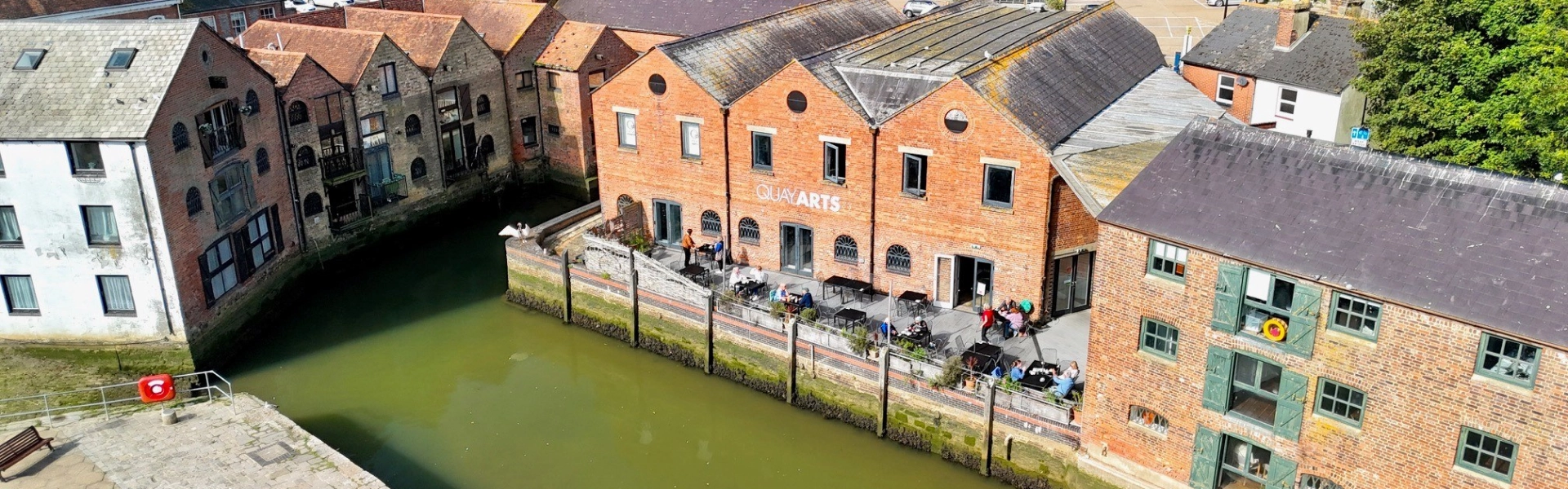 A canal lined with red brick warehouses and an outdoor cafe terrace; a QUAY ARTS brick building overlooks the greenish water, with townhouses and trees beyond.