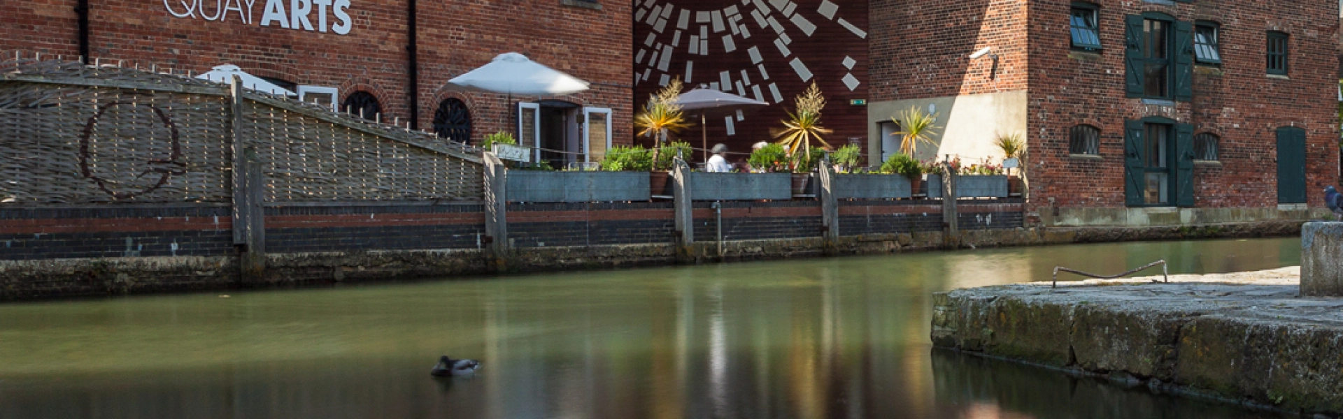 Red brick arts complex along a calm canal, featuring a QUAY ARTS sign, outdoor seating with a parasol, and a geometric mural reflected in the water.