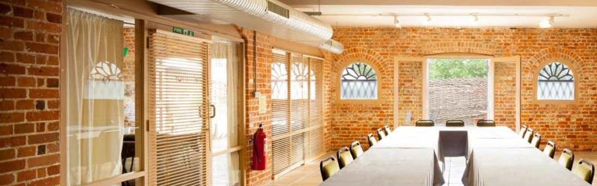 Industrial-style meeting room with brick walls, a long U-shaped grey-draped table, black chairs, exposed ducts, and large arched windows.