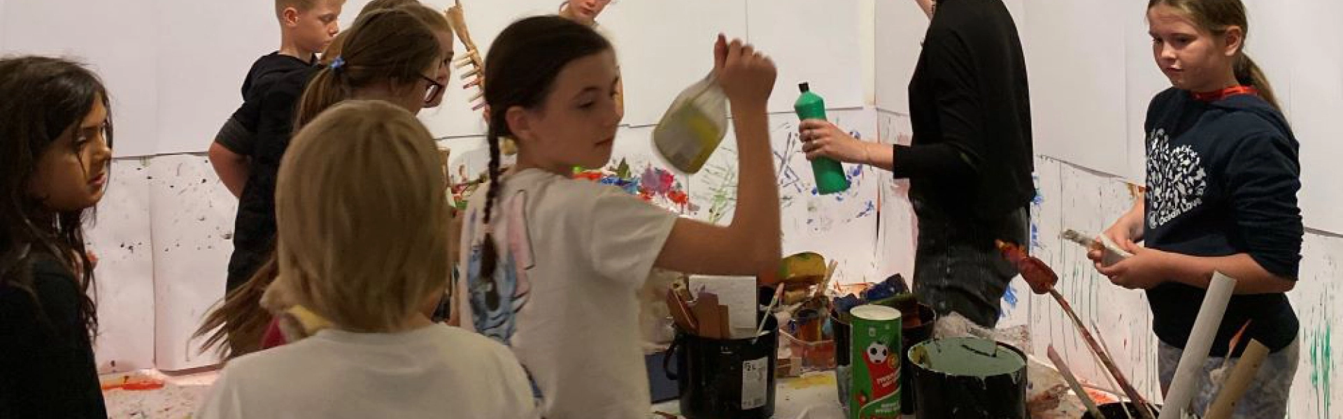 Group of children in a bright art room, painting on large white sheets taped to the walls; tables cluttered with paint, brushes and bottles.