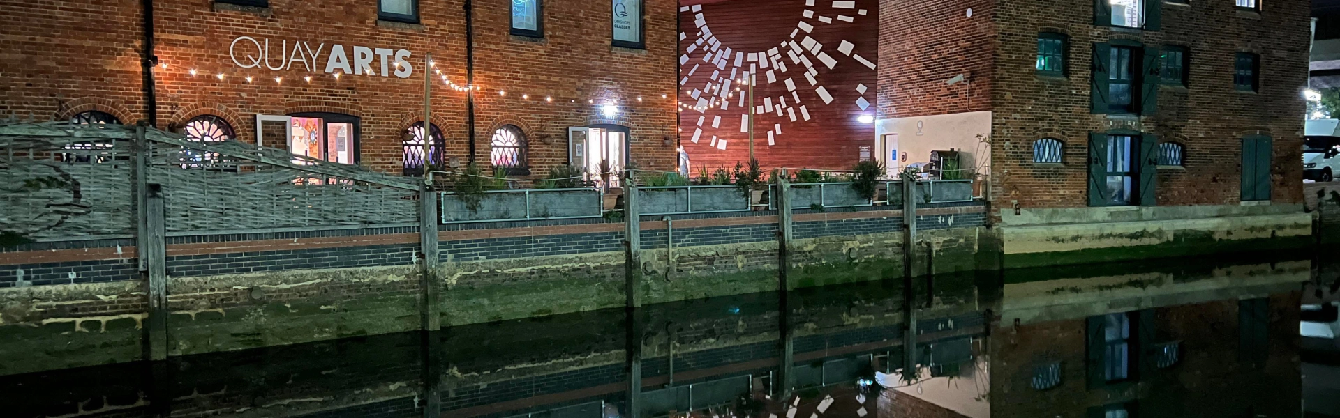 Night view of red brick Quay Arts building beside a canal, string lights, a mural of white panels on the red wall, and a reflected scene in the water.