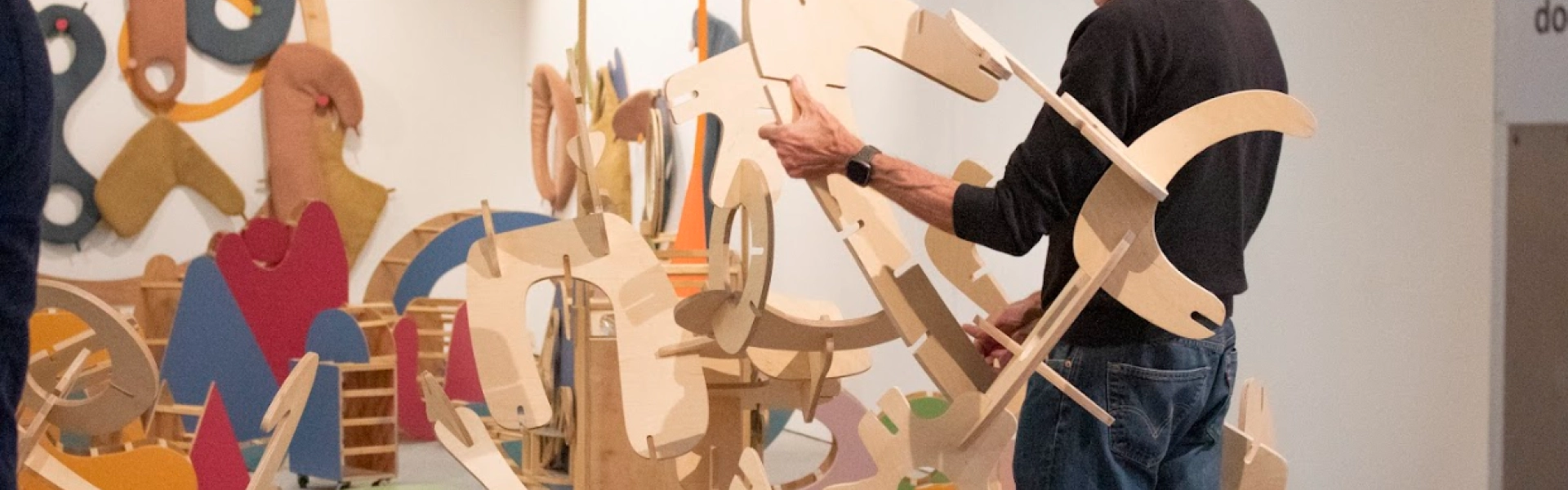 Person in a black top and jeans assembling a large wooden abstract sculpture, in a bright gallery filled with colourful wooden shapes and cutouts.