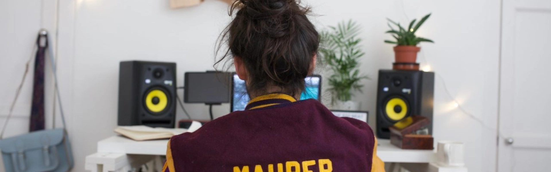 Back view of a person in a burgundy and yellow varsity jacket with 'MAURER' on it, seated at a desk with yellow speakers, plants and string lights.