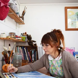A person with glasses and brown hair tied back sits at a cluttered art desk, painting with a brush on blue canvas, surrounded by jars and brushes.