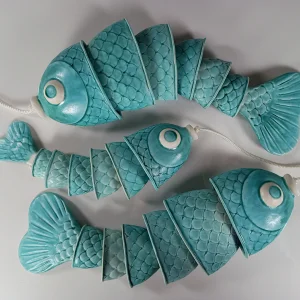 Three turquoise fish-shaped garlands with segmented, scale-patterned ceramic pieces strung on white cords against a light gray background.