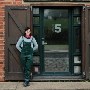 Person wearing green overalls and a gray sweater stands beside a green-framed glass door labeled Studio 5, with brown wooden shutters on a brick wall.