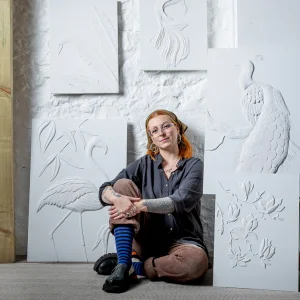 Person with orange hair, glasses, and tattoos sits cross-legged on a carpeted floor among white relief art boards of birds and plants against a white brick wall.