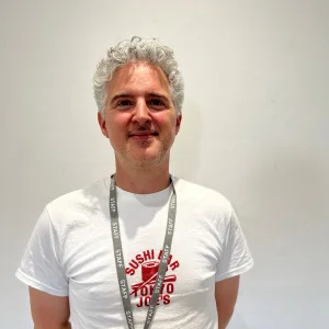 Smiling person with short grey curly hair wearing a white T-shirt with red print, a STAFF lanyard, standing against a light grey wall.