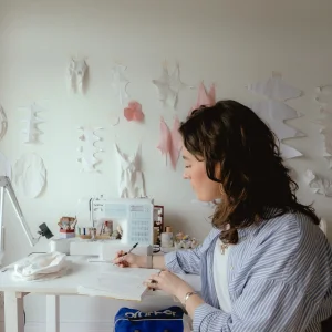 Side profile of a person with dark hair in a blue striped shirt, sketching at a white sewing desk with a Brother sewing machine; wall adorned with white paper shapes.