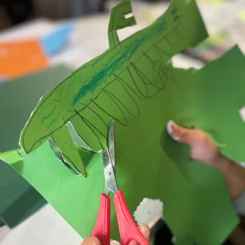 An image featuring a child's hands, they are cutting a green piece of card with scissors in their left hand. The card has a sea creature drawn on.