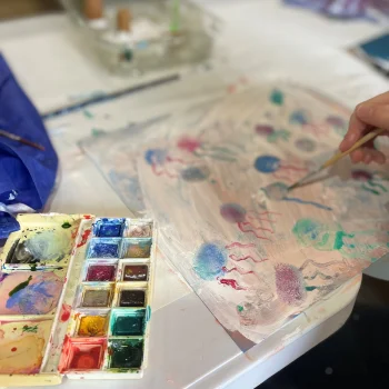 An image of a child's hands using watercolours to paint a series of colourful jellyfish on a piece of paper.