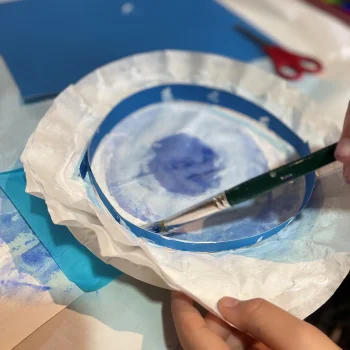An image of a child's hands, they are painting a piece of circular tissue paper blue to become a sea creature of their design.